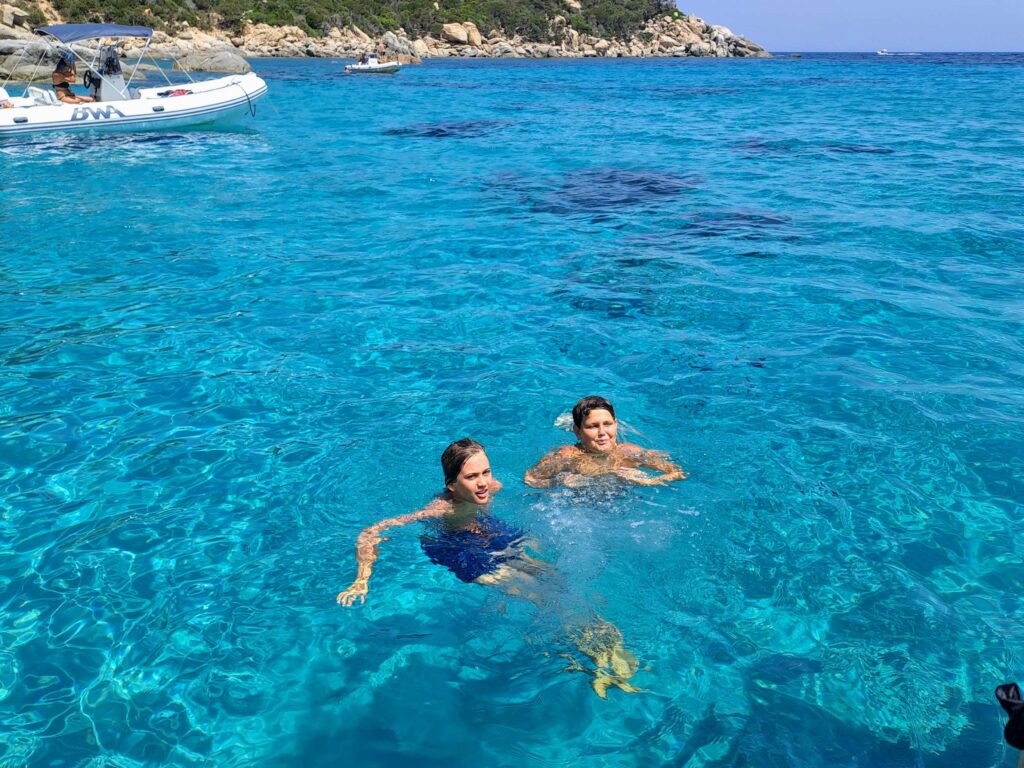 Two young people swimming and smiling in the strikingly clear turquoise waters of Mari Pintau, Sardinia, with a boat and coastline in the background.