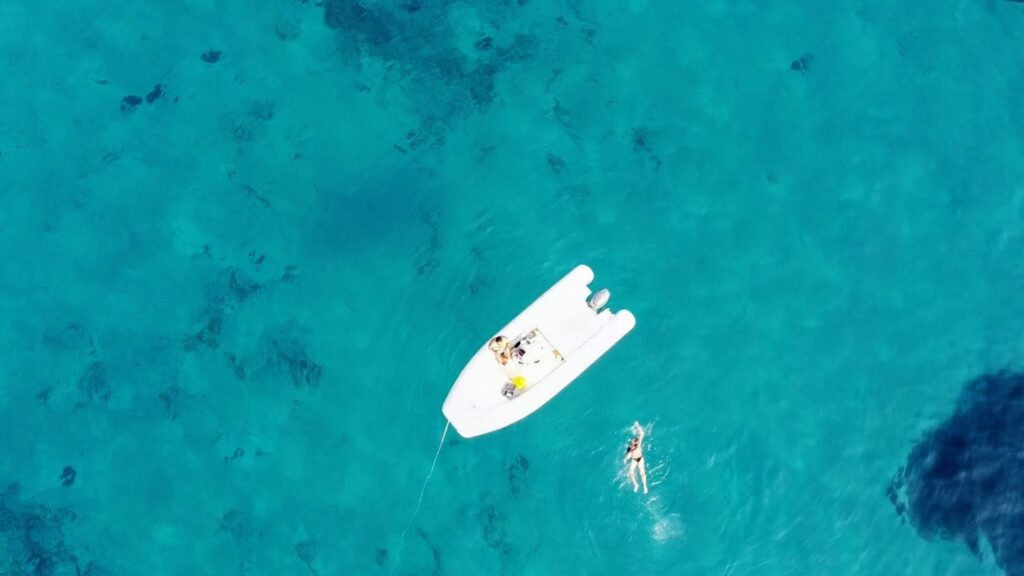 Aerial drone shot of a white inflatable boat and a person swimming in clear, vibrant turquoise water.