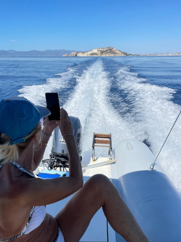 Woman in a bikini and cap taking a photo with her phone from the stern of a moving boat, showing the boat's wake and a distant island in the Gulf of Cagliari