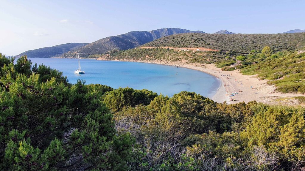 Elevated panoramic view of Mari Pintau beach, Sardinia, showing its crescent sandy shore, calm turquoise bay with a white sailboat, and surrounding green hills
